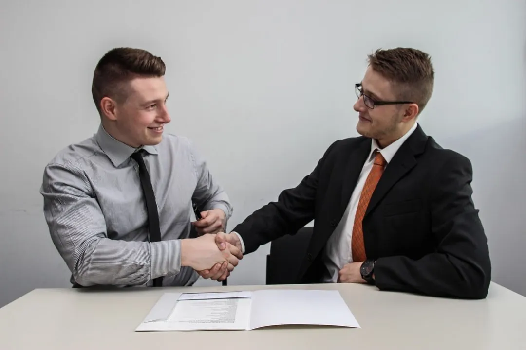 Two professionals shaking hands across a table with documents, representing serving as trustee.